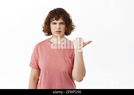 Confused girl pointing right at something strange, frowning and staring displeased or questioned, standing against white background in t-shirt Stock Photo