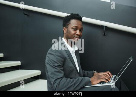 Graphic portrait of smiling African-American businessman using laptop while sitting on stairs in office hall Stock Photo