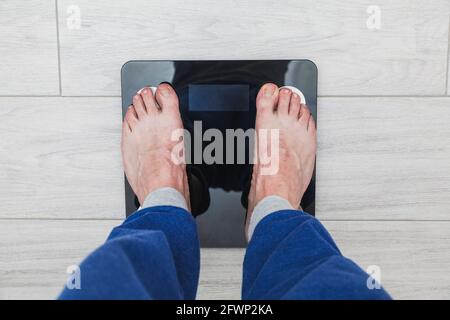 The feet of an unrecognizable man on top of an electronic scale where the weight is not visible. The man is wearing blue pajama pants and the floor is Stock Photo