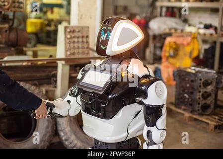 Humanoid robot on wheels with a monitor on his chest, shaking hands with a man. The old cluttered garage. Stock Photo