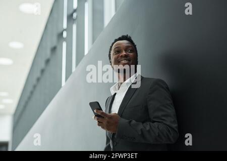 Graphic low angle portrait of carefree African-American businessman holding smartphone while standing against black background in office hall, copy sp Stock Photo
