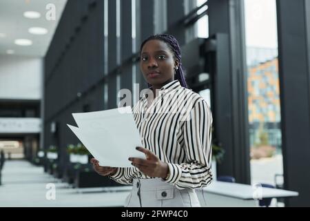 Graphic waist up portrait of confident African-American businesswoman looking at camera and holding documents while standing in office hall, copy spac Stock Photo