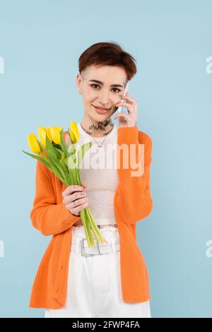 young tattooed woman in orange cardigan holding yellow tulips while talking on smartphone isolated on blue Stock Photo