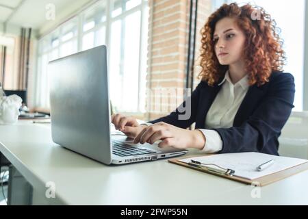 Beautiful young red-haired woman in a business suit works in an office with a laptop Stock Photo