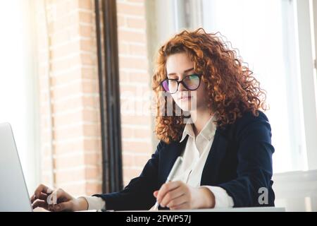 Beautiful young curly red-haired woman in a business suit and glasses works in the office on a laptop Stock Photo