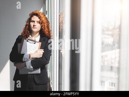 Portrait of a beautiful successful young business woman with red curly hair in the office interior stands near the window Stock Photo