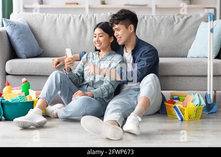 Loving chinese couple with cleaning tools resting, using smartphone Stock Photo