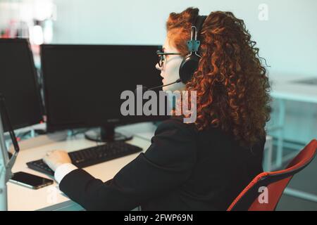 Portrait of a beautiful young woman operator with red curly hair and glasses in the interior of the call center office works at the computer and uses Stock Photo