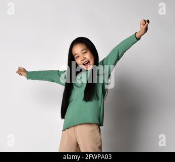 Happy screaming, singing loudly Asian kid girl in green hoodie and pants stands with arms up and outstretched Stock Photo