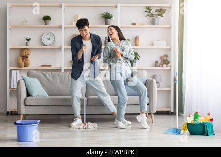 Funny asian couple singing songs while cleaning apartment, full length Stock Photo