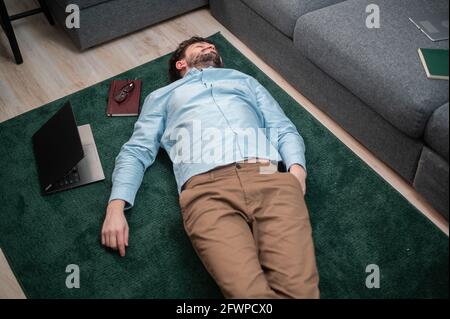In order not to hurt his back while working, the young man decided to relax and lay down on the floor next to the sofa. Stock Photo