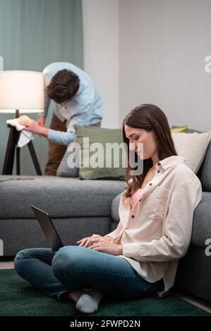 While the wife works remotely from home in a laptop, sitting on the floor, the husband repairs the floor lamp next to the sofa. Stock Photo