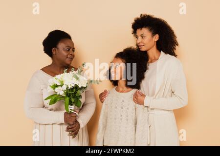 african american grandmother with bouquet of flowers looking at granddaughter and daughter on beige background Stock Photo
