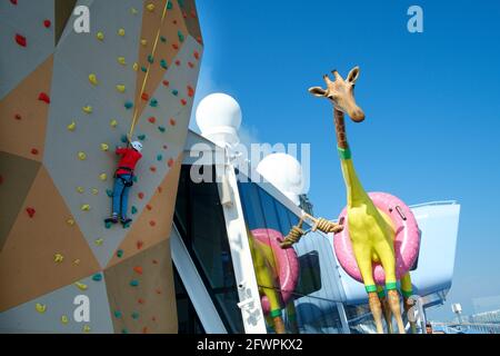 A young boy on the climbing wall next to a giant Giraffe called Gigi by french artist Jean Francois aboard Royal Caribbean ship Anthem of the Seas. Stock Photo