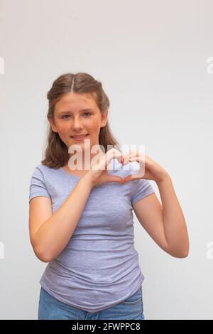 Portrait of a smiling young woman with freckled face showing heart gesture with two hands and looking at camera on light background Stock Photo