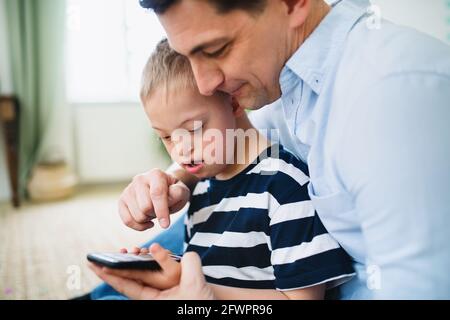 Father with happy down syndrome son indoors at home, using smartphone. Stock Photo