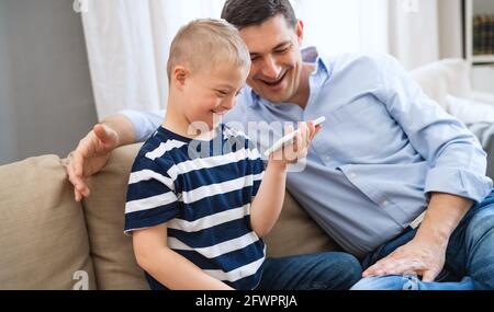 Father with happy down syndrome son indoors at home, using smartphone for watching. Stock Photo