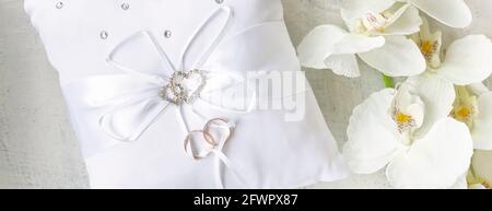 banner with white pillow for wedding rings with rings and rhinestone hearts near white orchid on a white textured background. Wedding or engagement co Stock Photo