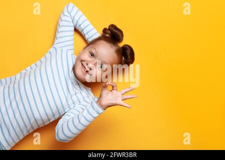 Top view of a cute adorable little girl with bundles of hair on a yellow background. The child shows the sign ok. Close up copy space. Stock Photo