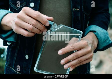 closeup of a young man, wearing a denim jacket and a casual green t-shirt, unscrewing the cap from a rectangular reusable water bottle Stock Photo