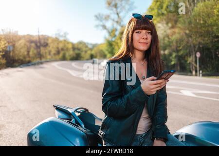 Motorcycle local travel. Portrait of smiling woman in a leather jacket and sunglasses sitting on a motorcycle and using a smartphone. World Motorcycli Stock Photo