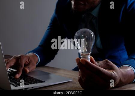 Mid section of businessman holding light bulb using laptop on wooden surface against grey background Stock Photo