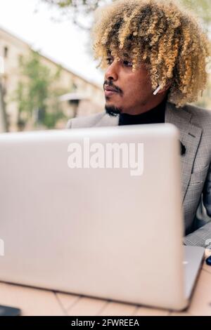 African business man with afro hair sitting in cafeteria and using his laptop. He is wearing earphones and listening to music. Stock Photo