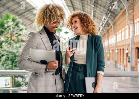 Happy afro business man wearing formal clothes talking with his coworker and using mobile phone. They are in Atocha train station, in Madrid. Stock Photo