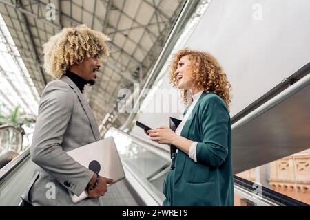 Business woman with formal wear using her mobile phone and talking with her african coworker in train station. Stock Photo
