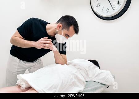 Male physiotherapist wearing face mask giving back massage to unrecognized woman lying in stretcher. Stock Photo