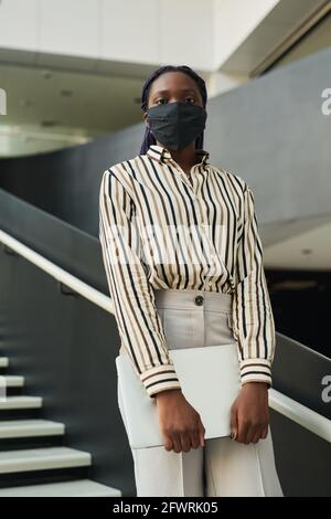 Vertical portrait of confident African-American businesswoman wearing mask and looking at camera while posing in office hall Stock Photo