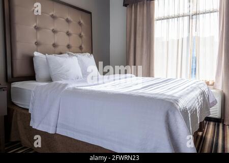 A spotless clean queen size hotel bed by a window in a bright hotel room. Upholstered pastel colored head board and ready made bed with white cover of Stock Photo