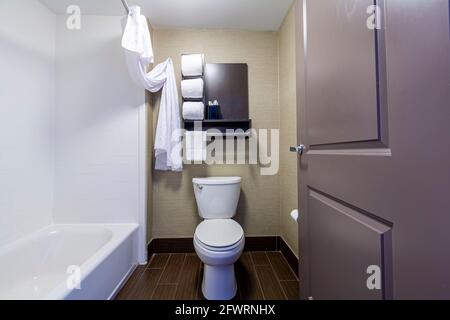 A small bathroom and toilet in a hotel with white towels folded and placed on shelves. A bathtub with shower curtain is on the left. A minimalistic de Stock Photo