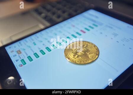A man holds a bitcoin coin in his hand. The graphs of changes in cryptocurrency rates are shown on the laptop monitor Stock Photo