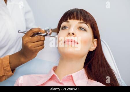 Closeup of face of beautiful young Caucasian woman receiving face oxygen peeling at cosmetology center. Red haired girl enjoying skin rejuvenation and revitalization. Needle free biorevitalization Stock Photo