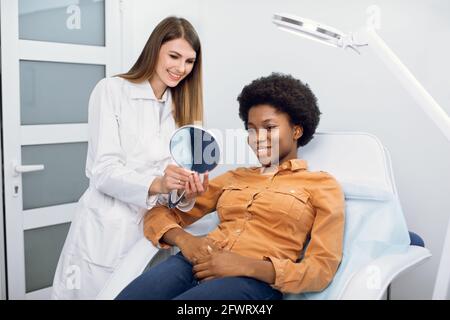 Consultation in cosmetology clinic. Female beauty doctor talking with patient, young African American woman, looking at the mirror and satisfied after the beauty procedures