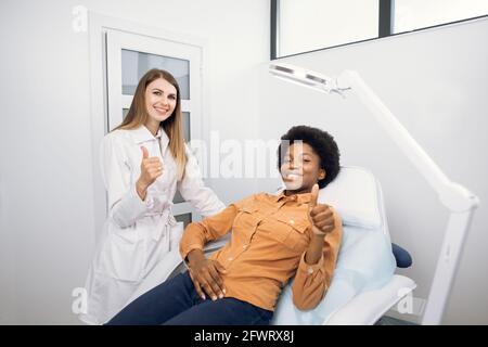 Happy young African American smiling woman, satisfied with results of cosmetic procedures in clinic of esthetic medicine, showing thumb up to camera together with her female doctor cosmetologist Stock Photo