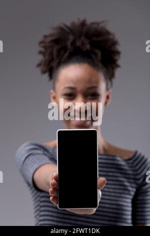 Happy young Black woman extending her blank mobile phone to the camera in a vertical position with focus to the smartphone on a grey studio background Stock Photo