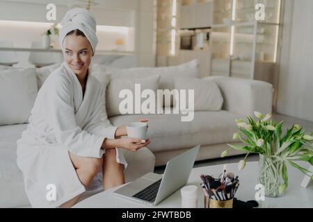 Young happy woman freelancer in bathrobe drinking tea and working remotely on laptop Stock Photo