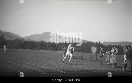 1951, historical, outside in a large field, on a grass track, senior school male competitiors taking part in a running race, watched by the event officials, some male and female teachers in the formal clothes of the era, Highlands, Scotland. A board with the runners names written in chalk can be seen standing on the grass. Stock Photo