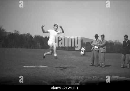 1951, outside in a large sports field and watched by two teachers in the formal clothes of the era, one wearing a hat and a fellow schoolboy, a young male in mid-air doing the long jump. A track and field event combing the need for speed, strength and agility in the attempt to leap as far as possible from a take off point. Speed on the run-up and a high leap at takeoff are fundamentals of success in this event. In the modern Olympics since 1896, the long jump is an ancient sport, with its origins coming from soliders in their training, having to jump obstacles such as streams and ravines. Stock Photo