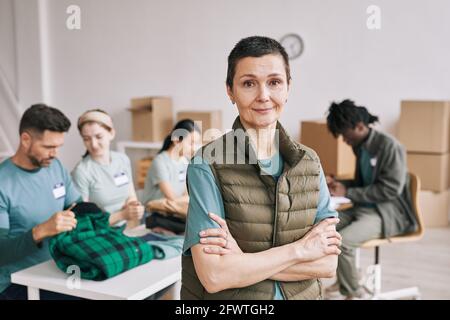 Waist up portrait of mature woman looking at camera during help and donations event with diverse team of volunteers in background, copy space Stock Photo