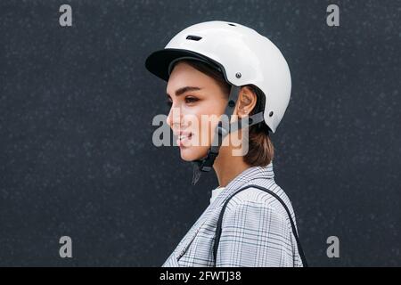 Side view of young woman wearing safety helmet. Female in scooter helmet standing at black wall. Stock Photo