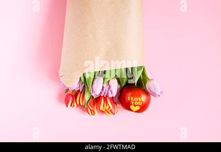 Bouquet of beautiful spring tulip flowers wrapped in craft paper bag and red apple with words I love You on soft pink background. Stock Photo