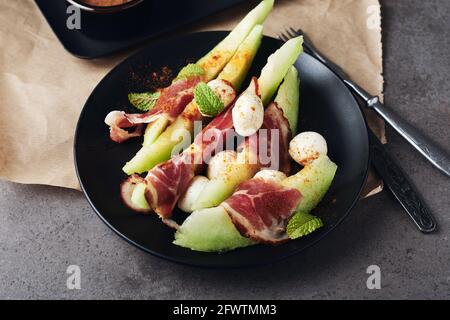 Melon with prosciutto, mozzarella balls and mint on a dark background. Stock Photo