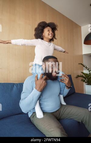 Black young father giving daughter piggyback ride when sitting on couch at home Stock Photo