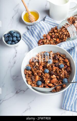Homemade granola, muesli with pieces of dark chocolate, nuts and blueberries in bowl on white marble background. Healthy breakfast. Stock Photo