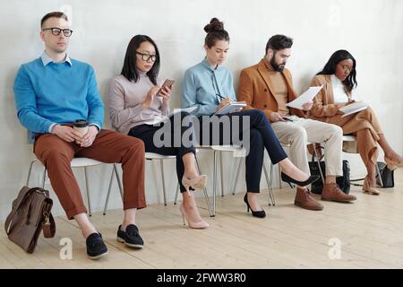 Group of serious young multi-ethnic job applicants in smart casual outfits sitting on chairs in line and preparing for interview in corridor Stock Photo