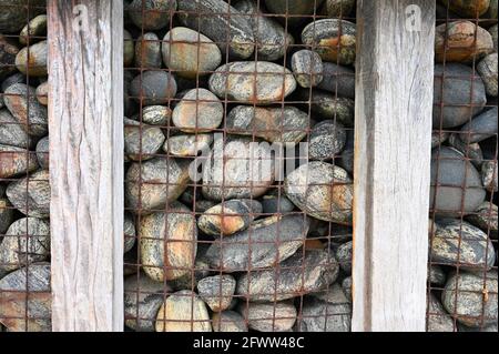 Abstract close up of outdoor retaining wall. Large stones behind rusted metal mesh with sunbleached wooden pillars. Isle of Lewis, Scotland. Stock Photo