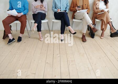 Legs of unrecognizable people using gadget and checking resumes while sitting in line Stock Photo